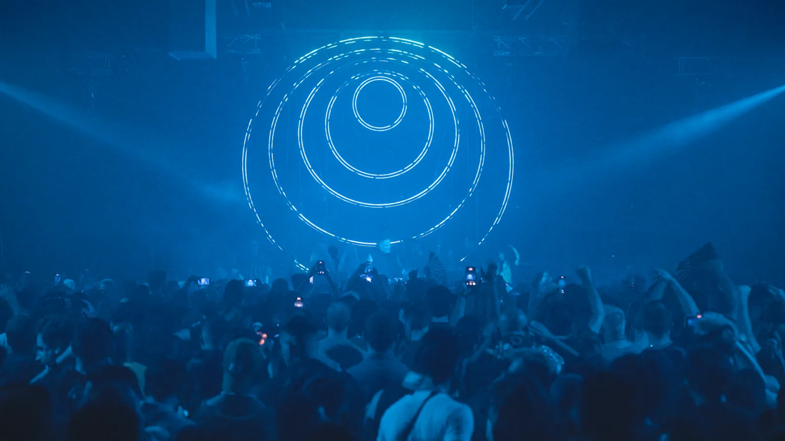 A packed Demo Room crowd with phones raised beneath concentric LED rings in blue light